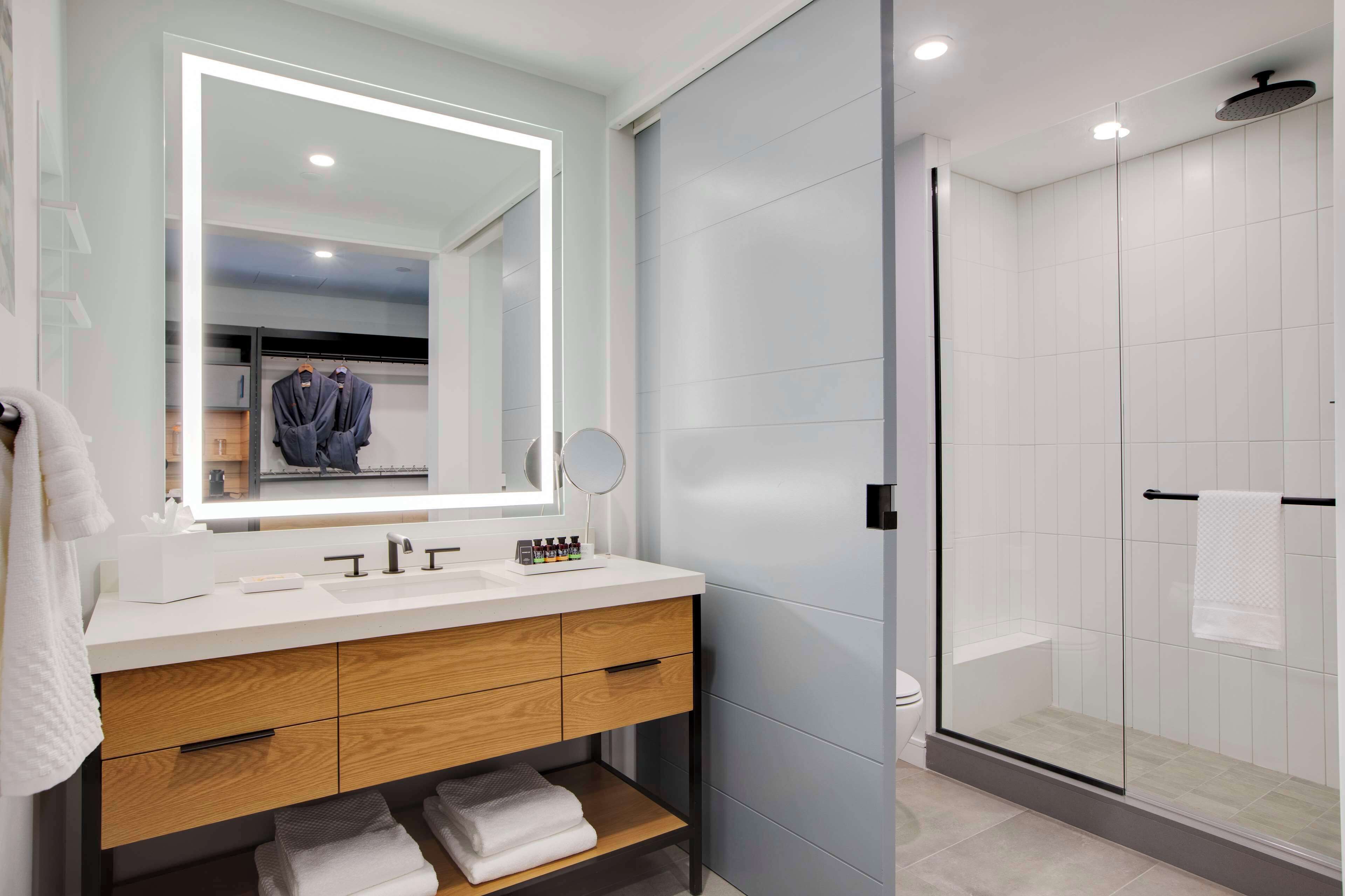 A modern bathroom with a white vanity, wooden drawers, a large mirror, and a glass-enclosed shower.