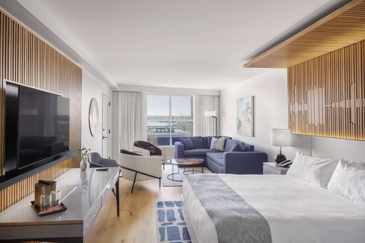 A modern hotel room with a large bed, a sofa, a TV, and a desk with a lamp.