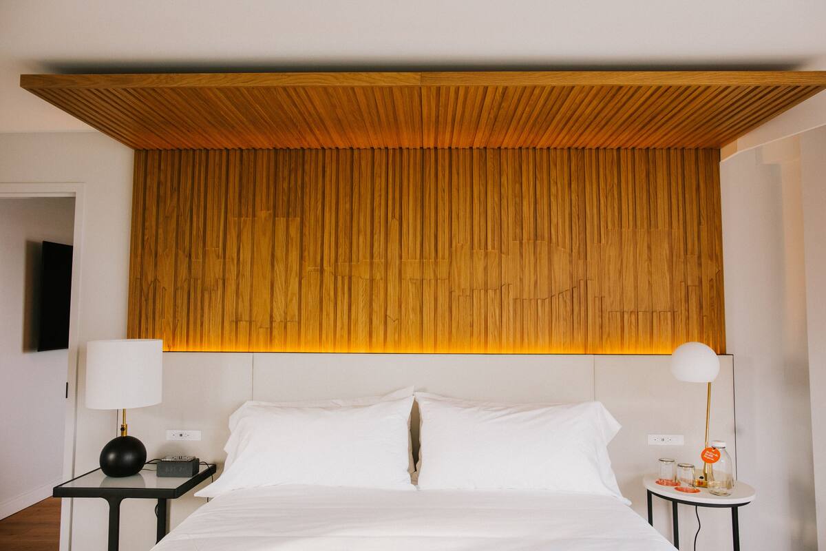 A hotel room with a wooden headboard, a bed with white linens, a nightstand with a lamp, and a small table with a lamp and a bottle.