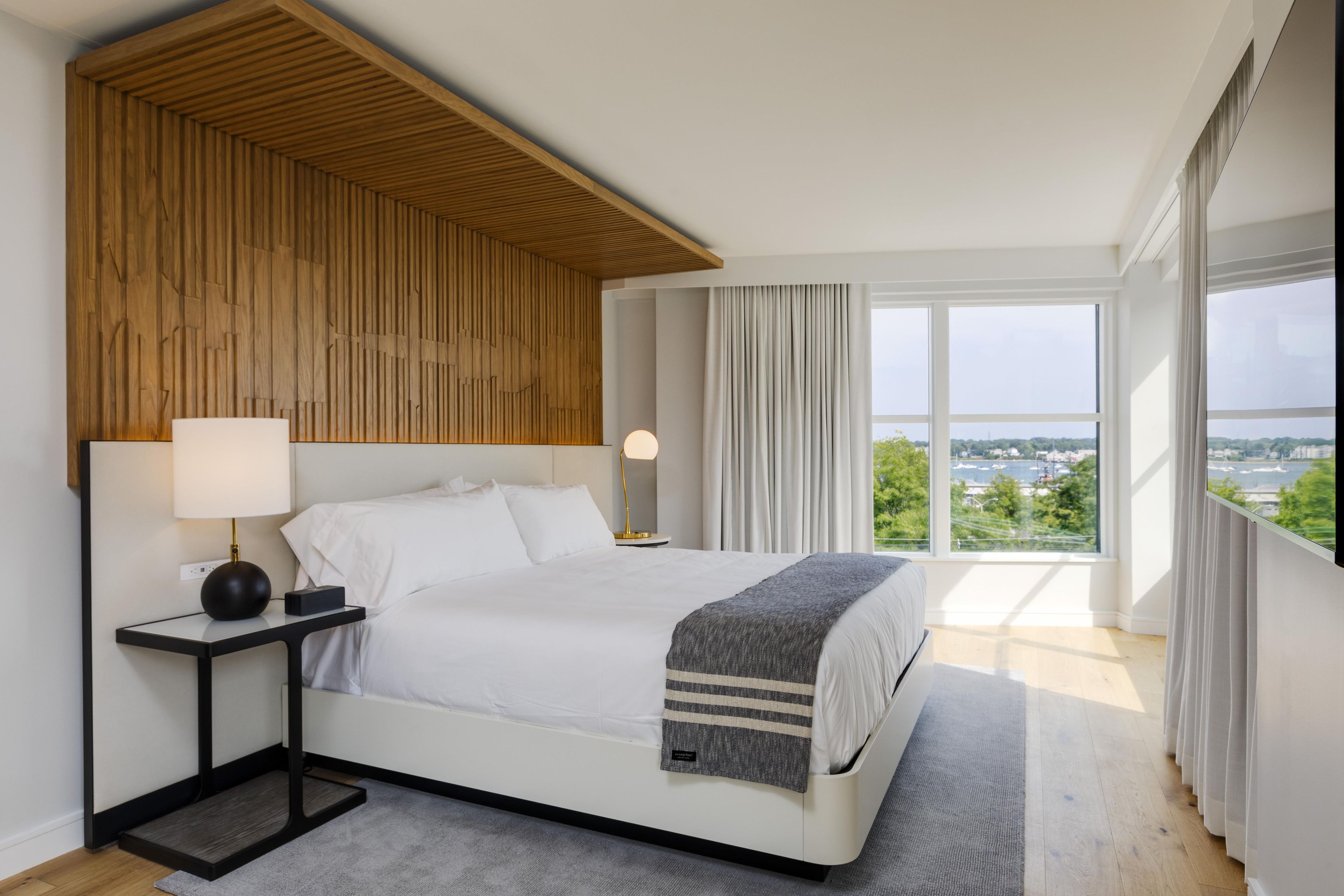 A modern bedroom with a bed, bedside tables, a view of the city through large windows, and a wooden ceiling design.