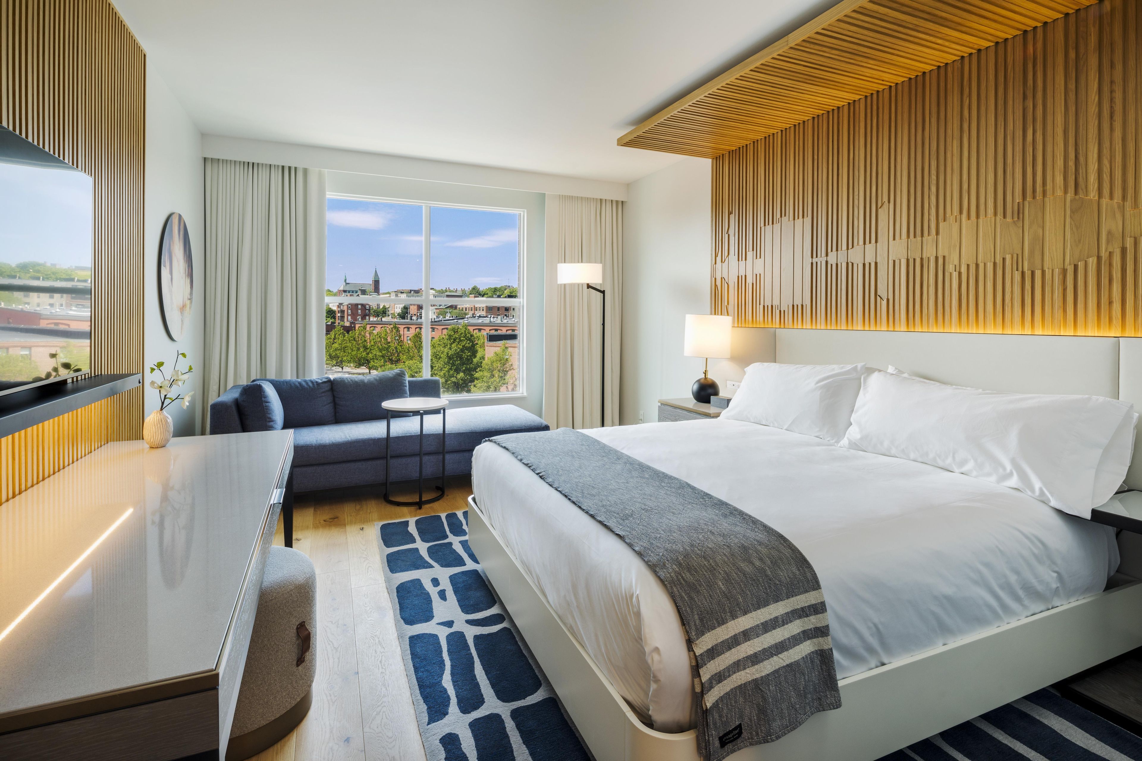 A hotel room with a bed, a desk, a chair, a small table, a lamp, and a view of the cityscape.