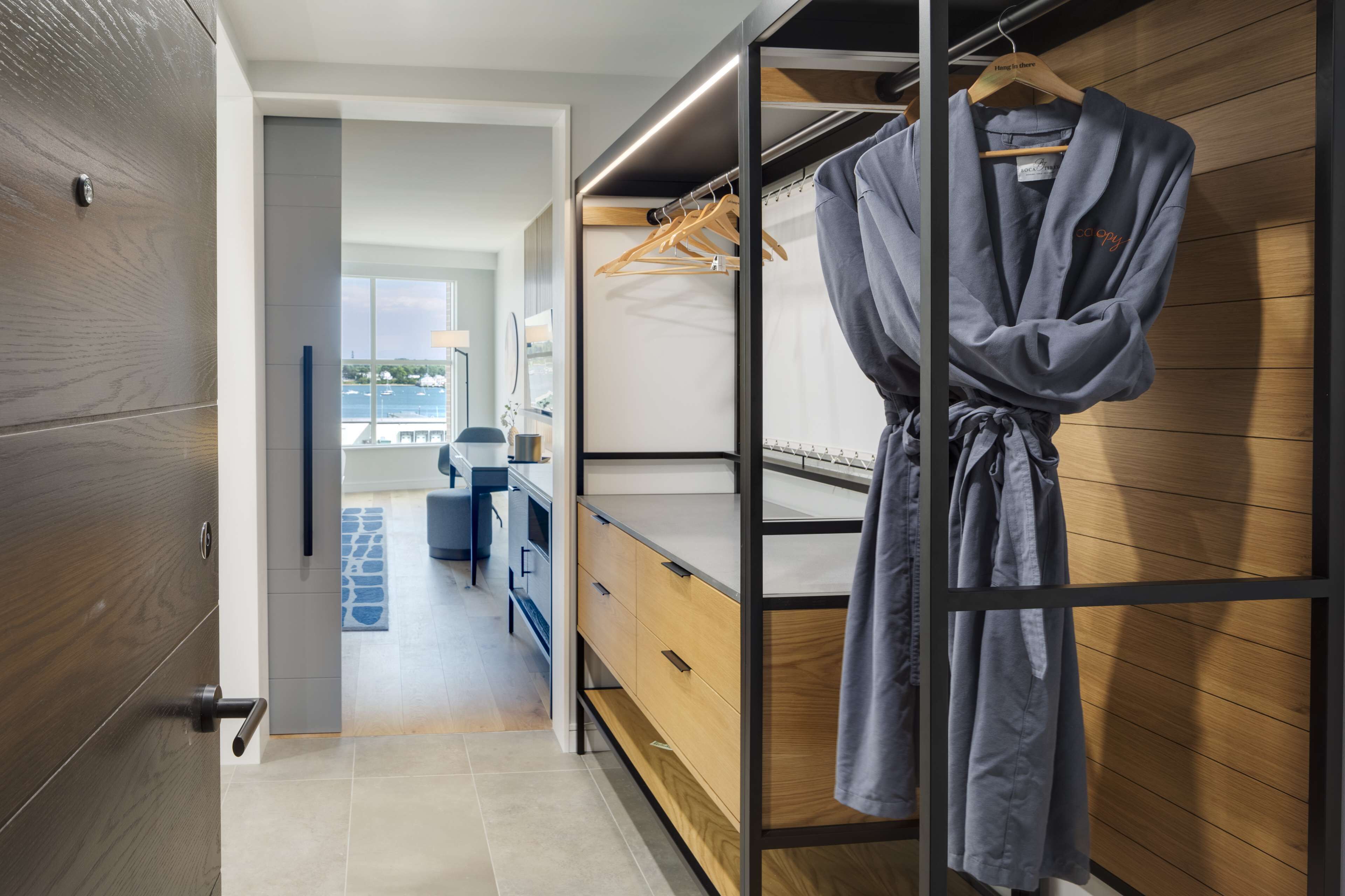 A modern bedroom with a wooden wardrobe, a hanging robe, and a view of a cityscape through a window.