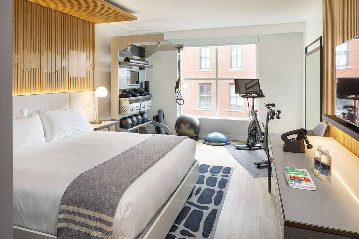 A hotel room with a bed, exercise equipment, a desk with a computer, and a window with a view of buildings.