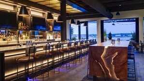 A modern bar with a long counter, multiple bar stools, and a large window offering a view of the cityscape. - Canopy by Hilton Portland Waterfront (Portland)