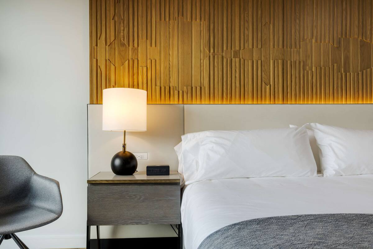 A hotel room with a bed, a nightstand with a lamp, a chair, and a wooden headboard.