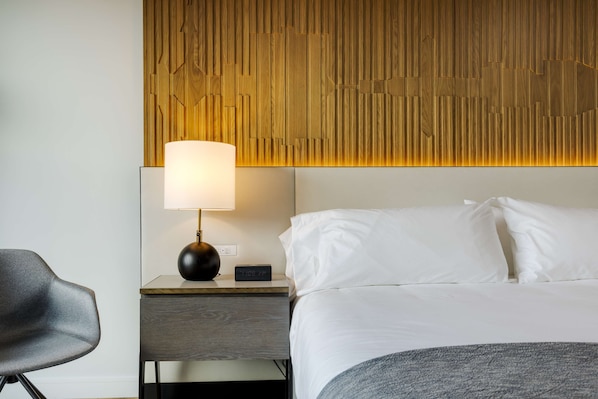 Una habitación de hotel con una cama, una mesita de noche con lámpara, una silla y un cabecero de madera.