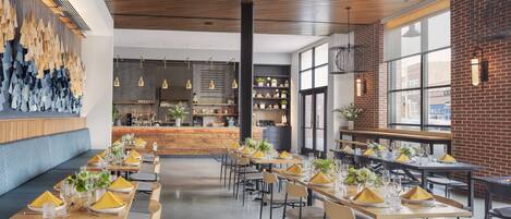 A modern restaurant with long wooden tables set for dining, a bar area, and a brick wall with large windows.