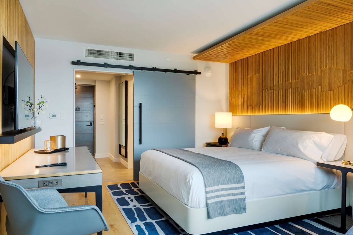 A modern hotel room with a bed, bedside table, lamp, desk, chair, and a TV.