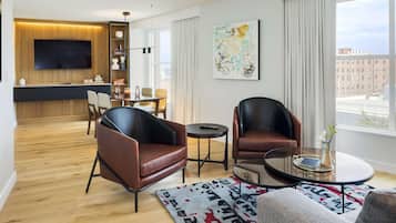 A modern living room with a flat-screen TV, leather armchairs, a glass coffee table, and a large window with a city view.