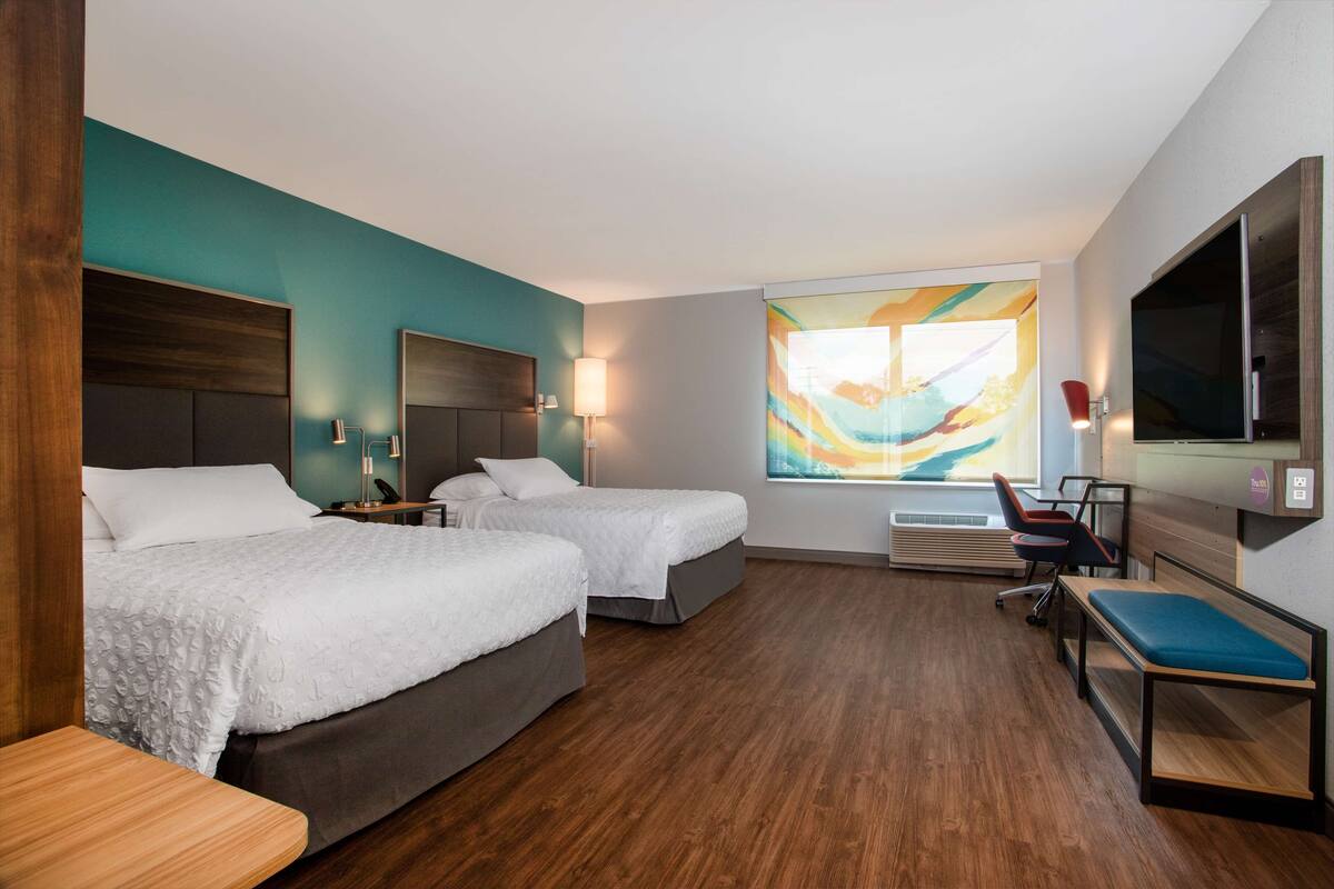 Ein Hotelzimmer mit zwei Betten, einem Schreibtisch, einem Stuhl, einem Fernseher und einem großen Fenster mit Wandgemälde.