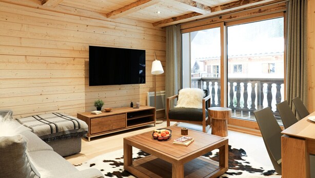 Een gezellige woonkamer met houten wanden en plafond, een flatscreen televisie aan de muur, een houten salontafel met fruit en een boek, een bank en een fauteuil.