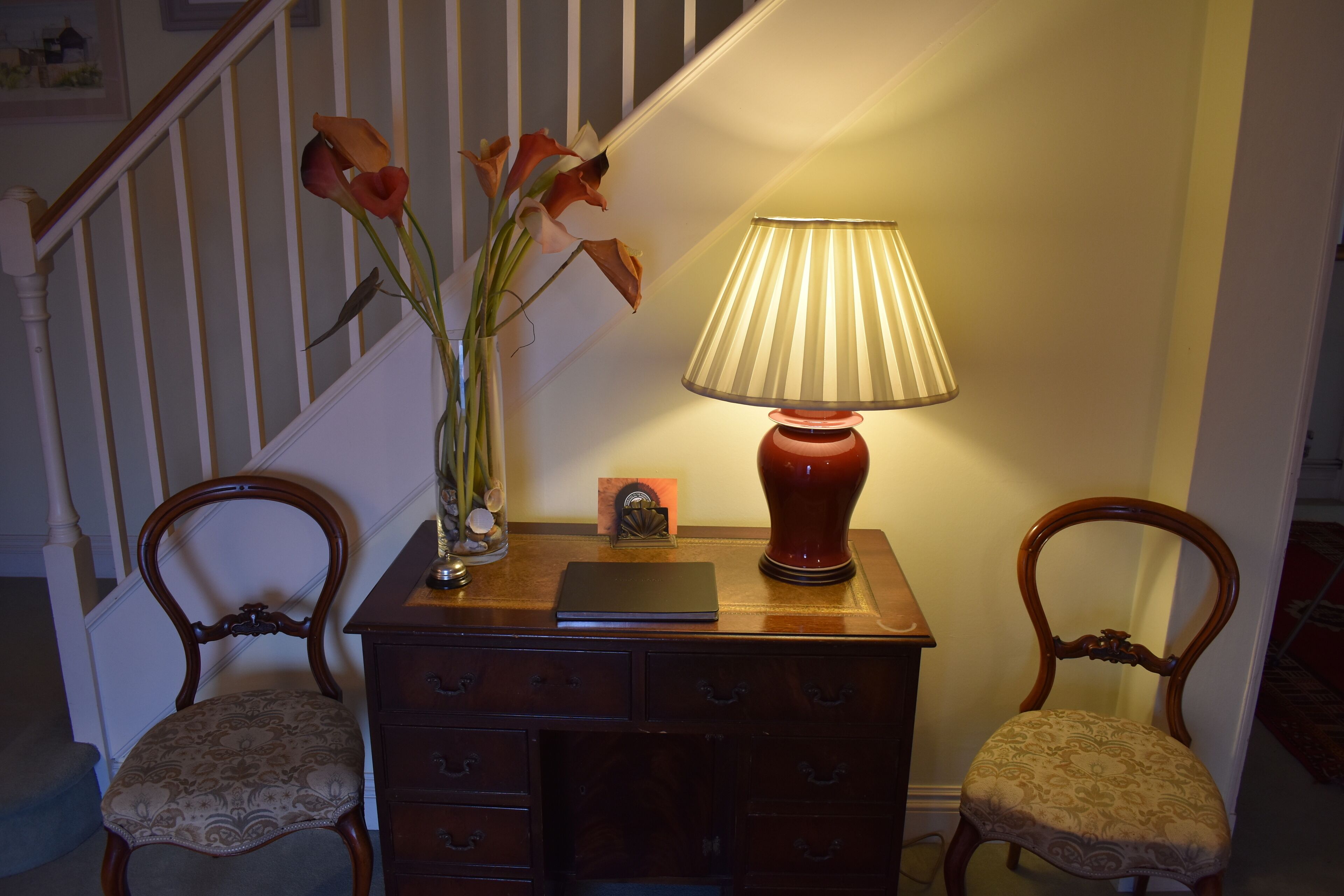 A wooden desk with a lamp, a vase with flowers, and two chairs in a room with a staircase in the background.