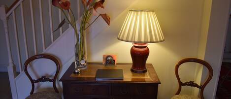 A wooden desk with a lamp, a vase with flowers, and two chairs in a room with a staircase in the background.