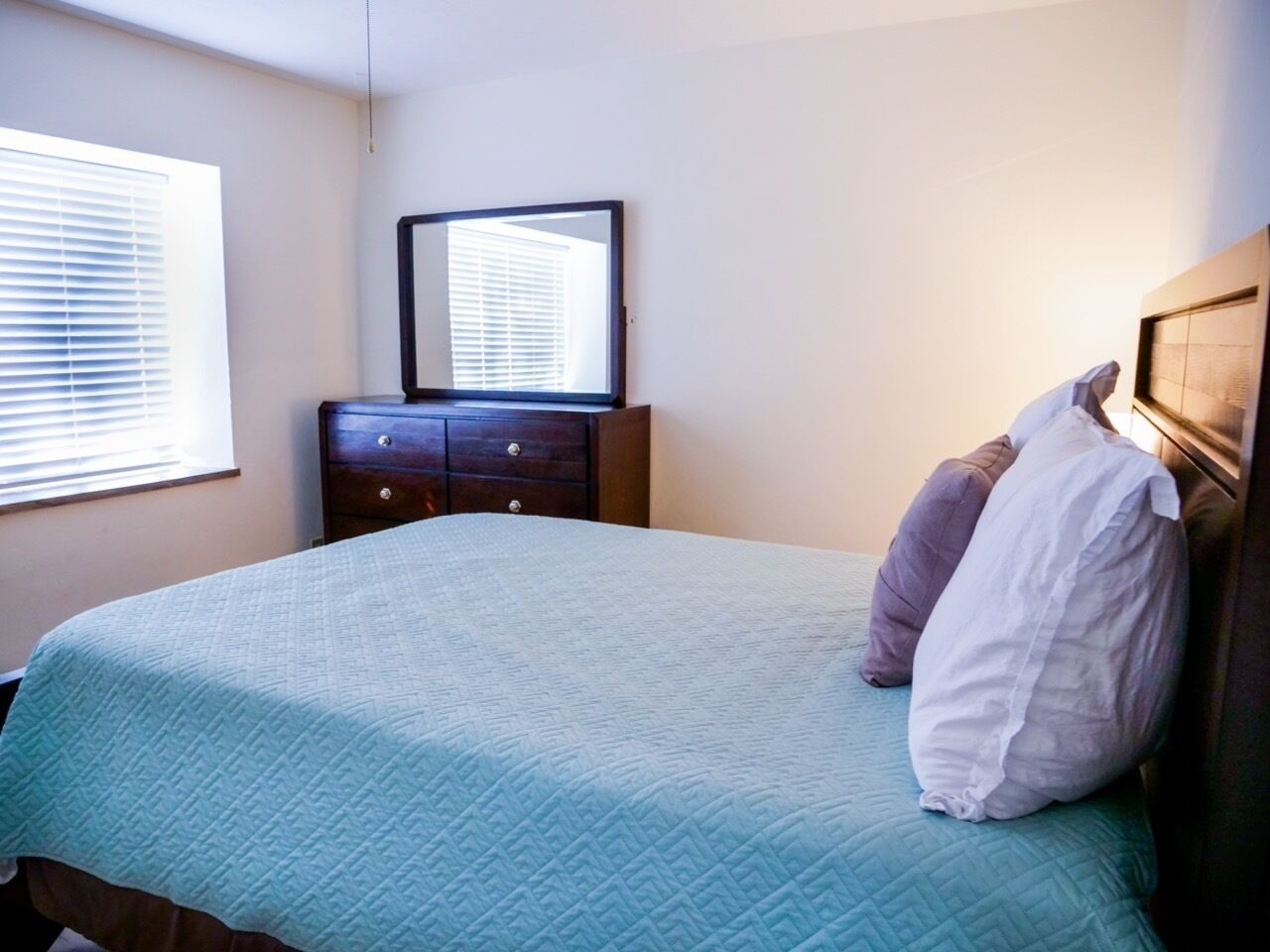 A bedroom with a bed, a dresser, a mirror, and a window with blinds.