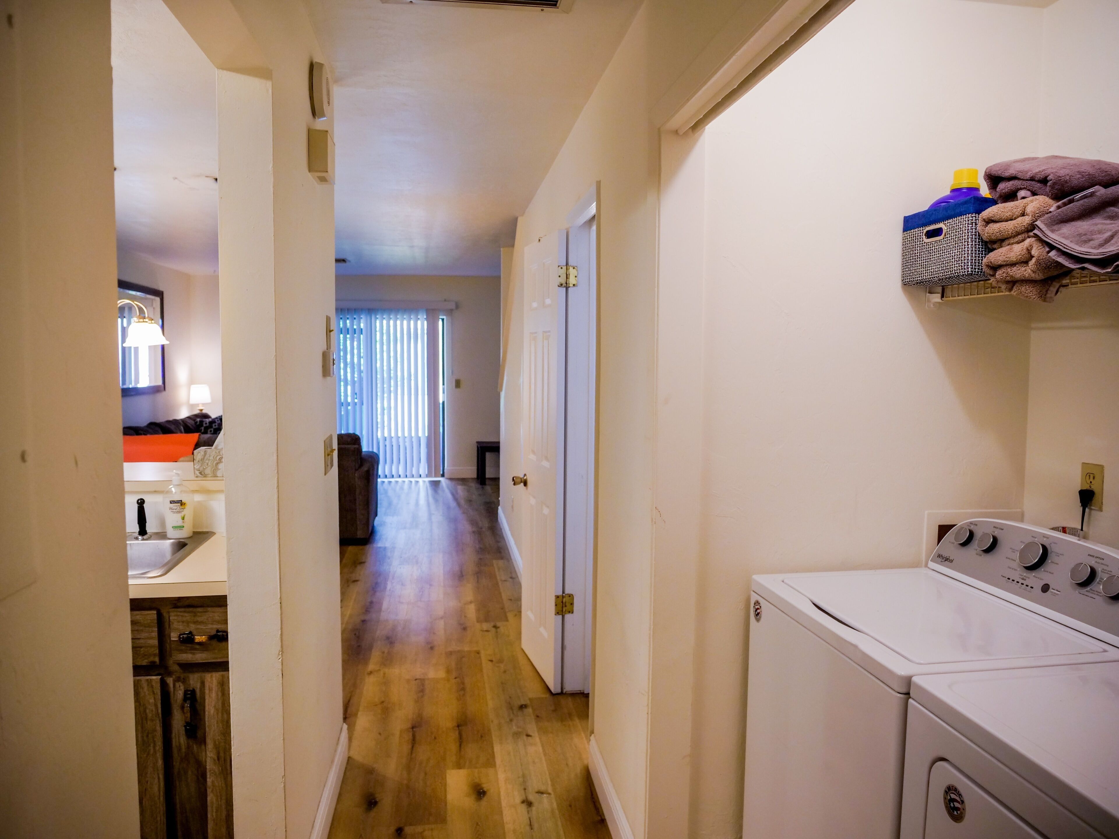 A laundry area with a washing machine, a drying rack with towels, and a hallway leading to a living room.