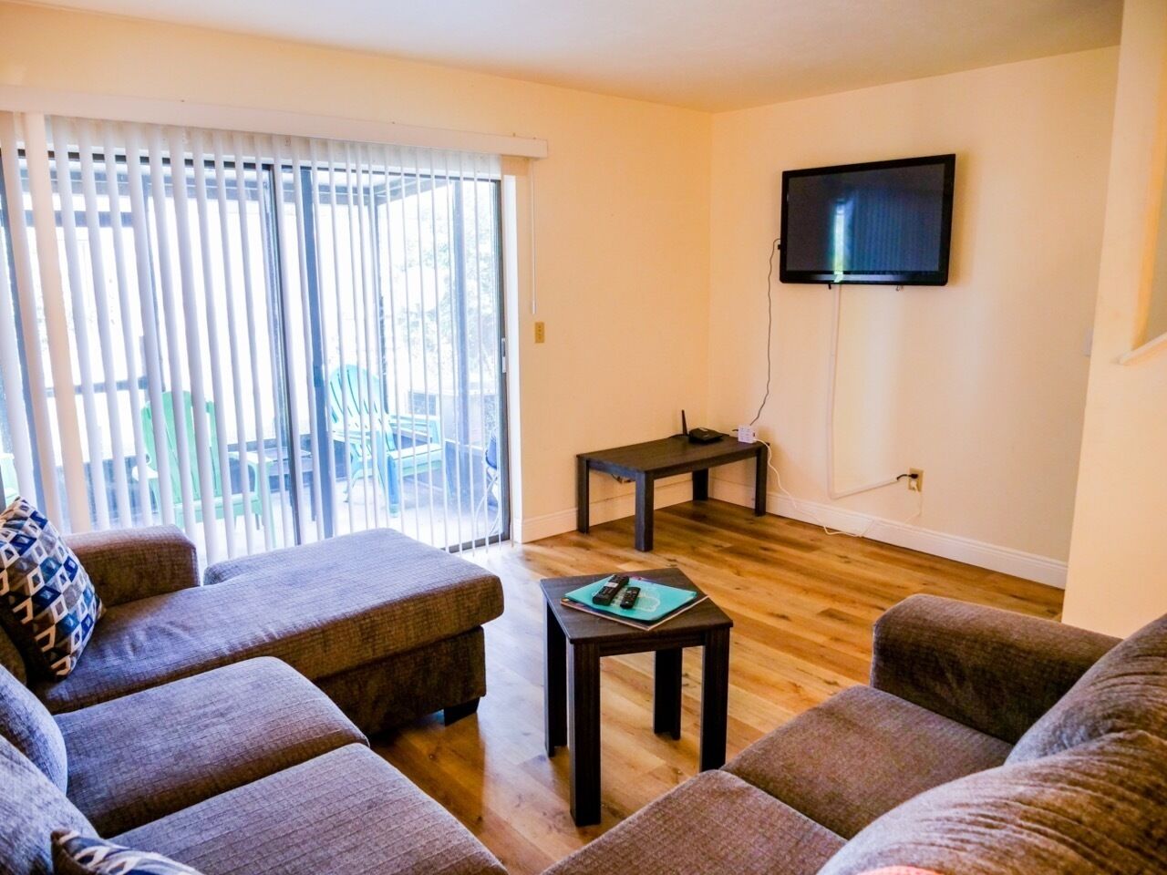A living room with a sofa set, a coffee table, a TV mounted on the wall, and a sliding glass door leading to an outdoor area.