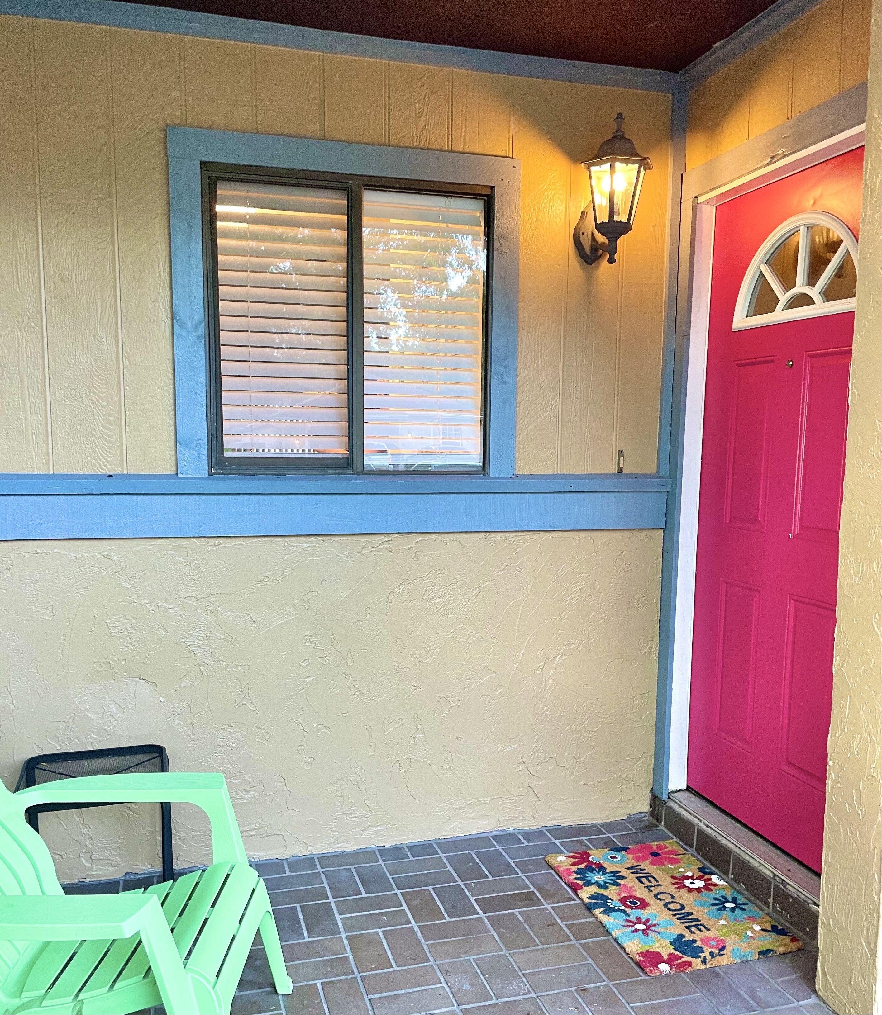 A front porch with a red door, a window with blinds, a green chair, and a welcome mat.