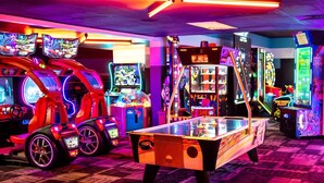 Une salle d’arcade lumineuse, avec différents jeux vidéo, notamment des jeux de course et de combat, et une table de billard au centre.