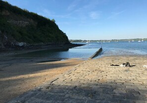 A beach with a stone pathway leading to the water, boats in the distance, and a cliff with vegetation.