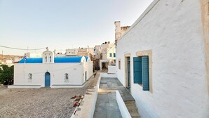 A narrow street with white buildings and blue doors, leading to a small courtyard with a stone floor and a few people sitting.