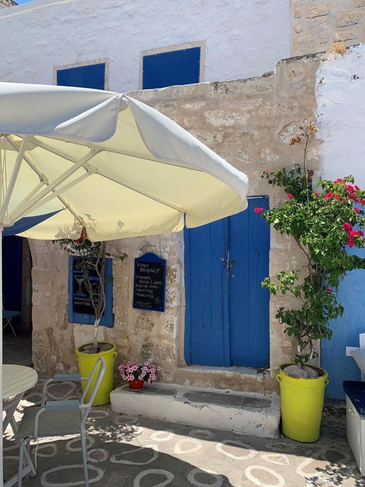 A quaint outdoor seating area with a table and chairs under a white umbrella, a blue door, and a signboard advertising breakfast options.