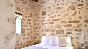 A rustic stone-walled bedroom with a wooden ceiling, a bed with white linens, and a small bedside table with a framed picture.