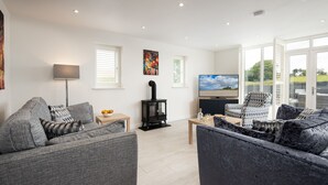 A modern living room with grey sofas, a black wood-burning stove, a flat-screen TV, and large windows offering a view of the outdoors. - Hot Tub Haven, sleeps 8 in rural scenic Northumberland (Chatton)