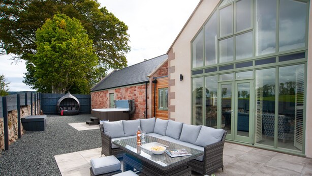An outdoor patio area with a glass-top table, grey cushioned seating, and a barbecue grill. - Hot Tub Haven, sleeps 8 in rural scenic Northumberland (Chatton)