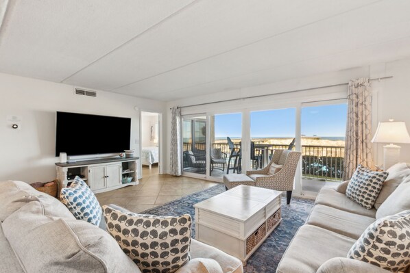 A living room with a flat-screen TV, a sofa set, a coffee table, and a view of the sea through large windows.