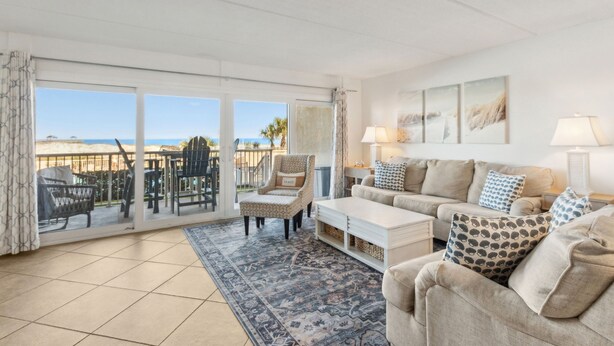 A spacious living room with a large sliding glass door leading to a balcony, a beige sofa set, a patterned rug, and a view of the sea. - Direct Pool and Oceanfront Views | Ground Level Amelia By The Sea | Exclusive Access to the Pier (Fernandina Beach)