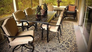 Une terrasse couverte avec une table et des chaises, entourée de grandes fenêtres donnant sur une zone boisée.