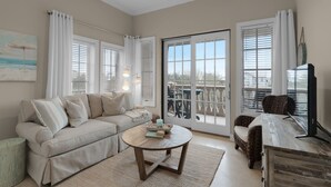 Un salon avec un canapé beige, une table basse ronde et un fauteuil en osier. De grandes fenêtres, ornées de rideaux blancs, offrent une vue sur l’extérieur.