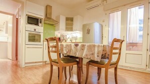 Un comedor con una mesa lista para cuatro personas, rodeada de sillas de madera. Se observa una cocina con armarios blancos y un refrigerador al fondo.