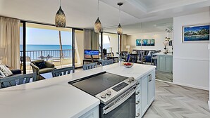 A modern kitchen with an island, large windows with a view of the sea, and a dining area with a view of the ocean. - Where the Pacific Stretches Forever Kihei Remodeled Oceanfront Condo Pool (Kihei)