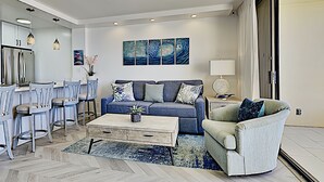 A modern living room with a sofa, coffee table, and chairs. There is a kitchen area with bar stools and a refrigerator. A large window provides natural light. - Where the Pacific Stretches Forever Kihei Remodeled Oceanfront Condo Pool (Kihei)