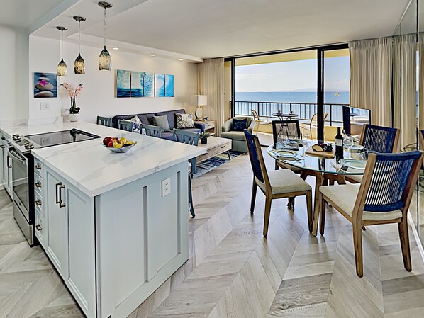 A modern kitchen with a central island, dining area with chairs, and a view of the sea through large windows.