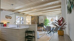Una cucina moderna con soffitto in legno, una zona pranzo con tavolo e sedie, e un angolo soggiorno con televisore e piante.