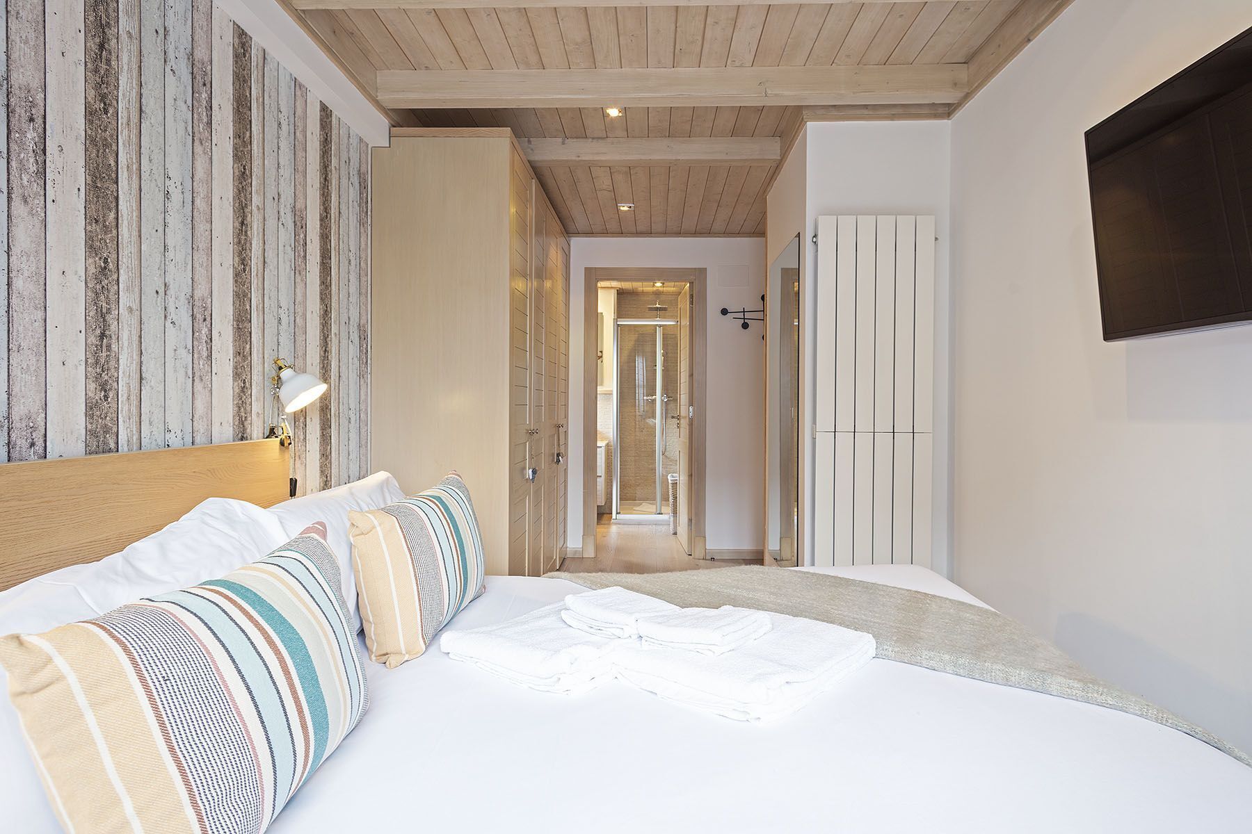 A bedroom with a bed, a bedside lamp, a wooden ceiling, a wardrobe, and a TV mounted on the wall.