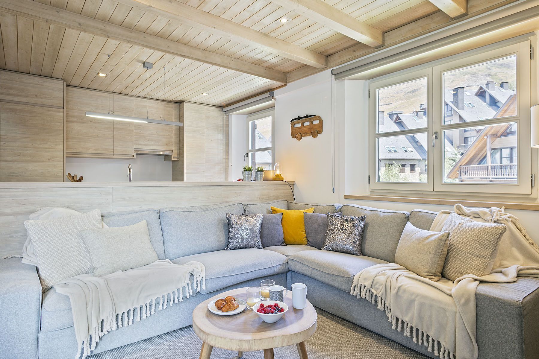 A modern living room with a light-colored sectional sofa, a round coffee table with breakfast items, and a large window with a view of buildings.