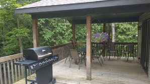 A covered outdoor deck with a grill, chairs, and a hanging flower arrangement. - Cardinal Crest of Sister Bay (Sister Bay)