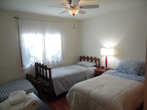 Quarto com duas camas de solteiro, uma cadeira de madeira, um ventilador de teto e uma mesa de cabeceira com abajur.