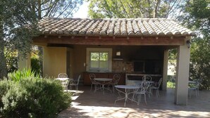 Eine überdachte Terrasse im Freien mit Ziegeldach, Grill und weißen Metalltischen und -stühlen.