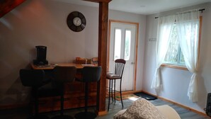 A cozy room with a wooden table, bar stools, a patterned sofa, a clock on the wall, and a window with curtains.