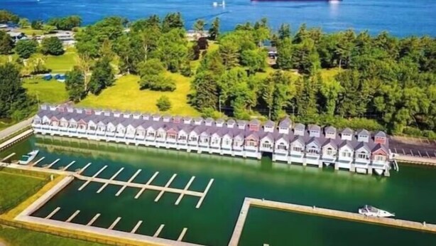 A marina with a row of docked boats, a large ship in the background, and a building with a row of small houses. - Gorgeous Townhouse & Boathouse in Cozy Carnegie Bay (Alexandria Bay)