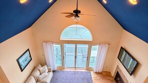 A high-ceilinged room with a ceiling fan, a large window, a sofa, a TV, and a fireplace. - Gorgeous Townhouse & Boathouse in Cozy Carnegie Bay (Alexandria Bay)