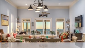 A spacious living room with a large window overlooking the ocean, a dining area with a table set for four, and a cozy seating area with a sofa and armchairs. - Two Palms | Your Gulf-Front Happy Place. Pets Welcome (Dauphin Island)