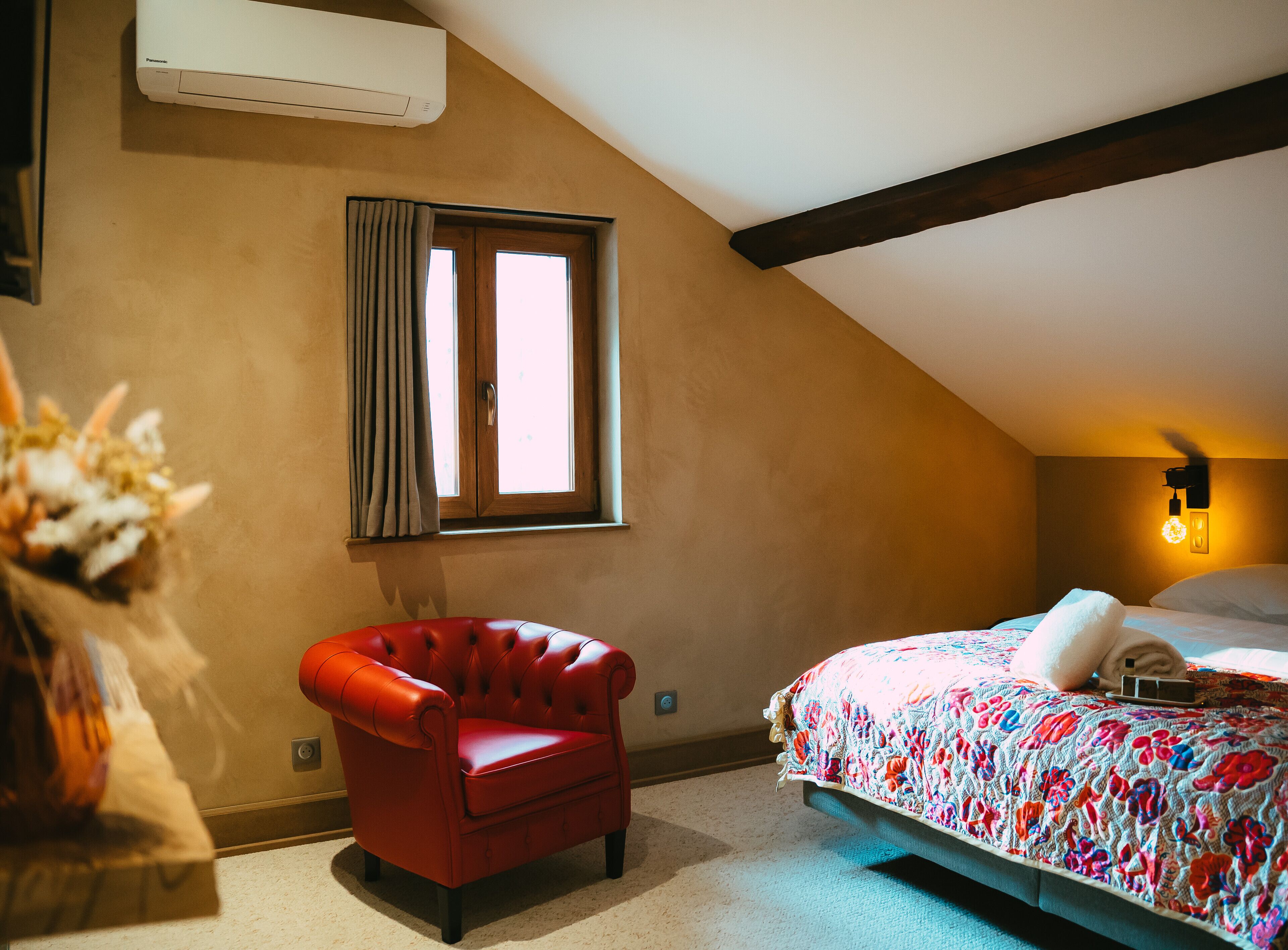 A bedroom with a sloped ceiling, a red leather chair, a bed with a floral bedspread, and a window with curtains.