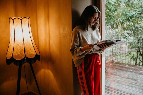 A person reading a book near a lamp and a window with a view of greenery.