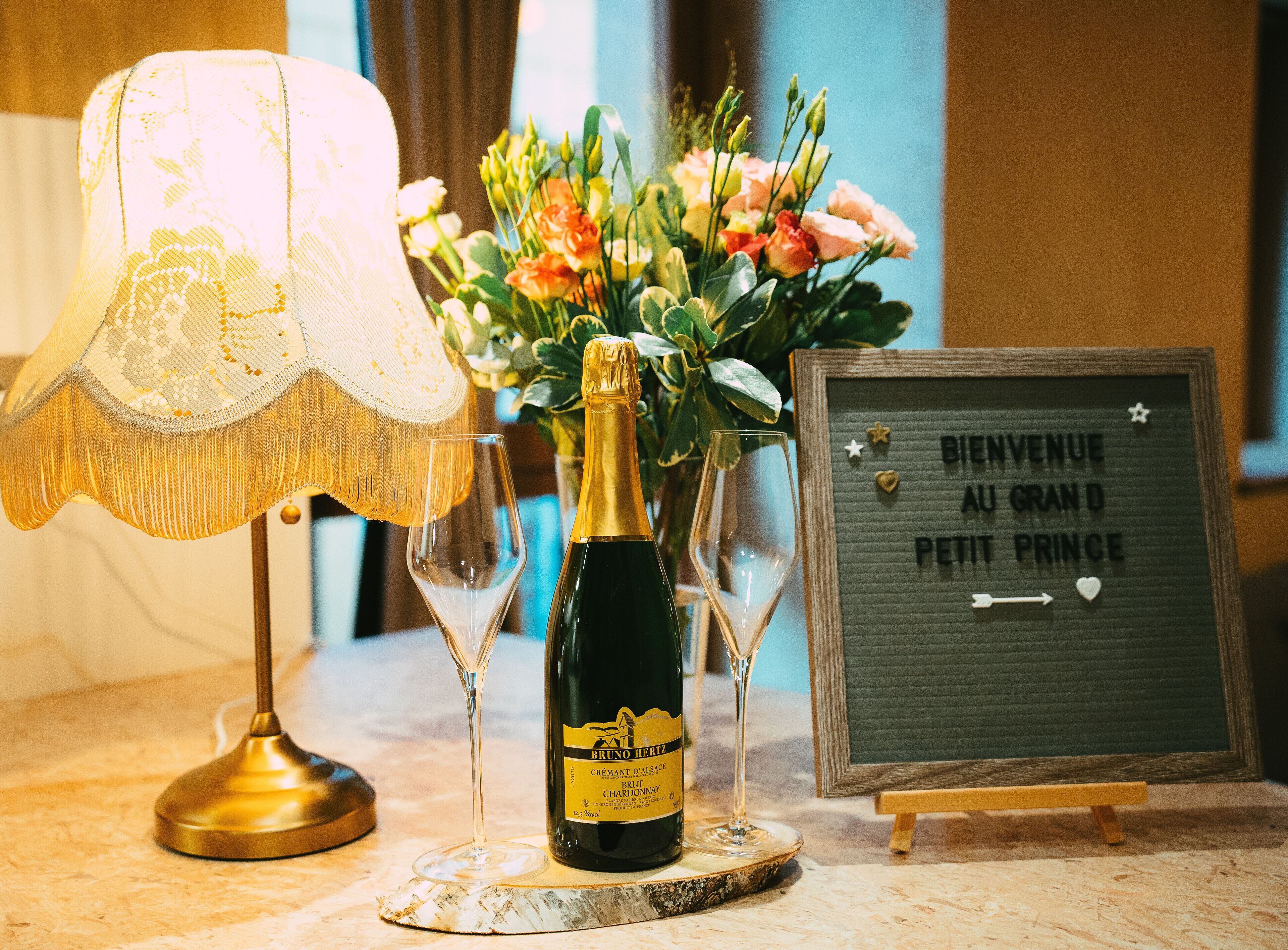 A bottle of Brut Chardonnay, two wine glasses, a decorative lamp, a floral arrangement, and a welcome sign.