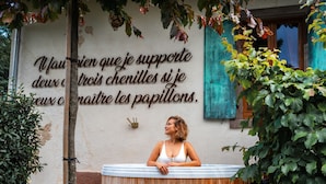 Une femme se détend dans un bain à remous en bois, et un proverbe français est inscrit sur le mur derrière elle.
